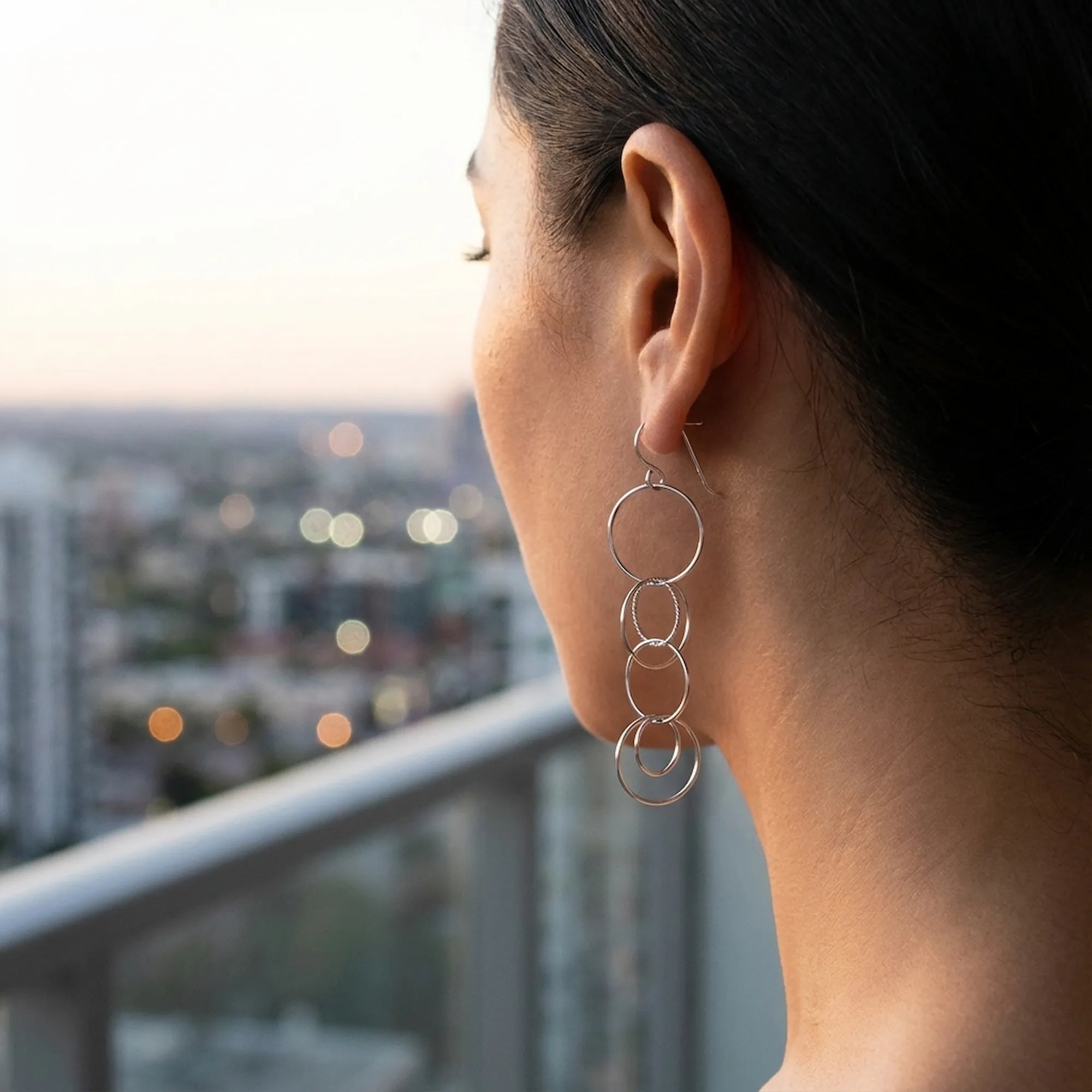 Close-up editorial photograph of a healthy earlobe comfortably wearing elegant, long interlocking silver circle drop earrings against a blurred city skyline at dusk, illustrating the comfort and luxury of hypoallergenic jewelry.
