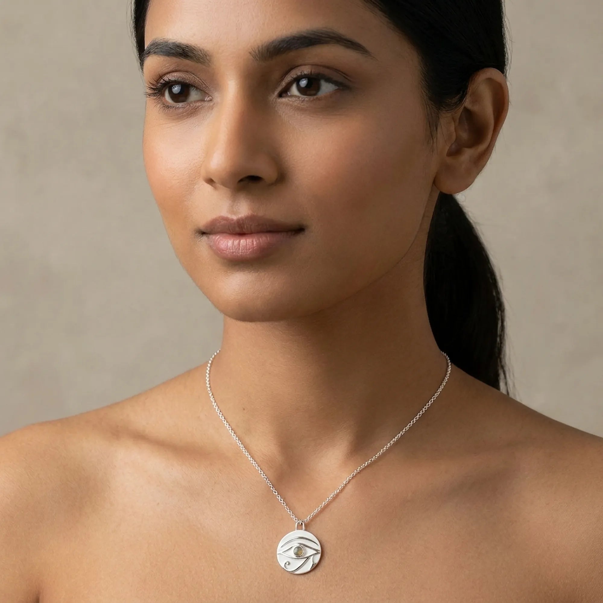 Woman wearing a silver necklace wedjat eye of horus pendant against a neutral background