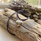 Heart-shaped locket pendant on a dark blue cord lying on a wooden surface with lavender and stones in the background.