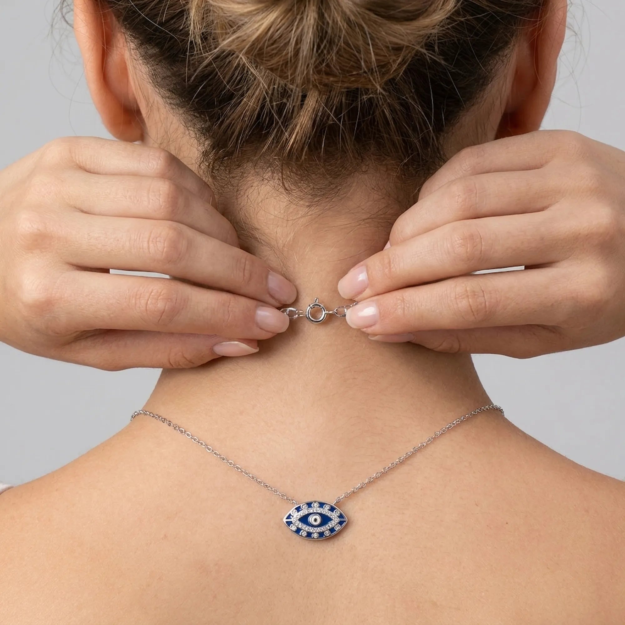 Model wearing a silver necklace with an eye pendant, adjusting closer clasp on a neutral background
