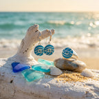 Blue earrings on a wooden block with seashells and beach sand in the background