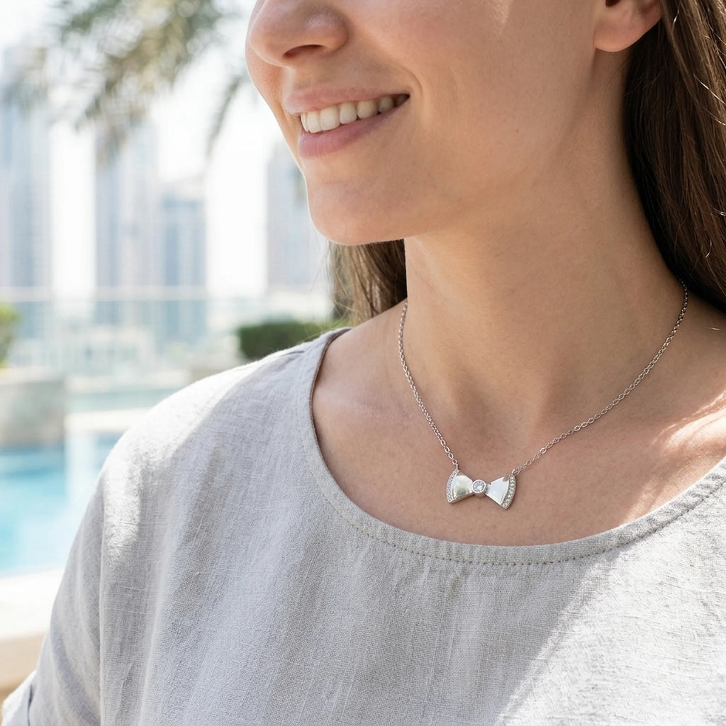 Woman wearing a silver bow necklace near a pool with a view of the Dubai Marina skyline blurred in the background.