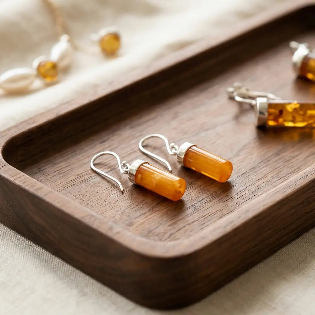 Pair of handmade amber cylinder earrings with silver caps resting on a dark walnut wood jewelry tray with a soft linen background