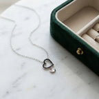 Black pavé open-heart necklace with polished silver crown charm on pale marble beside a green jewellery case.