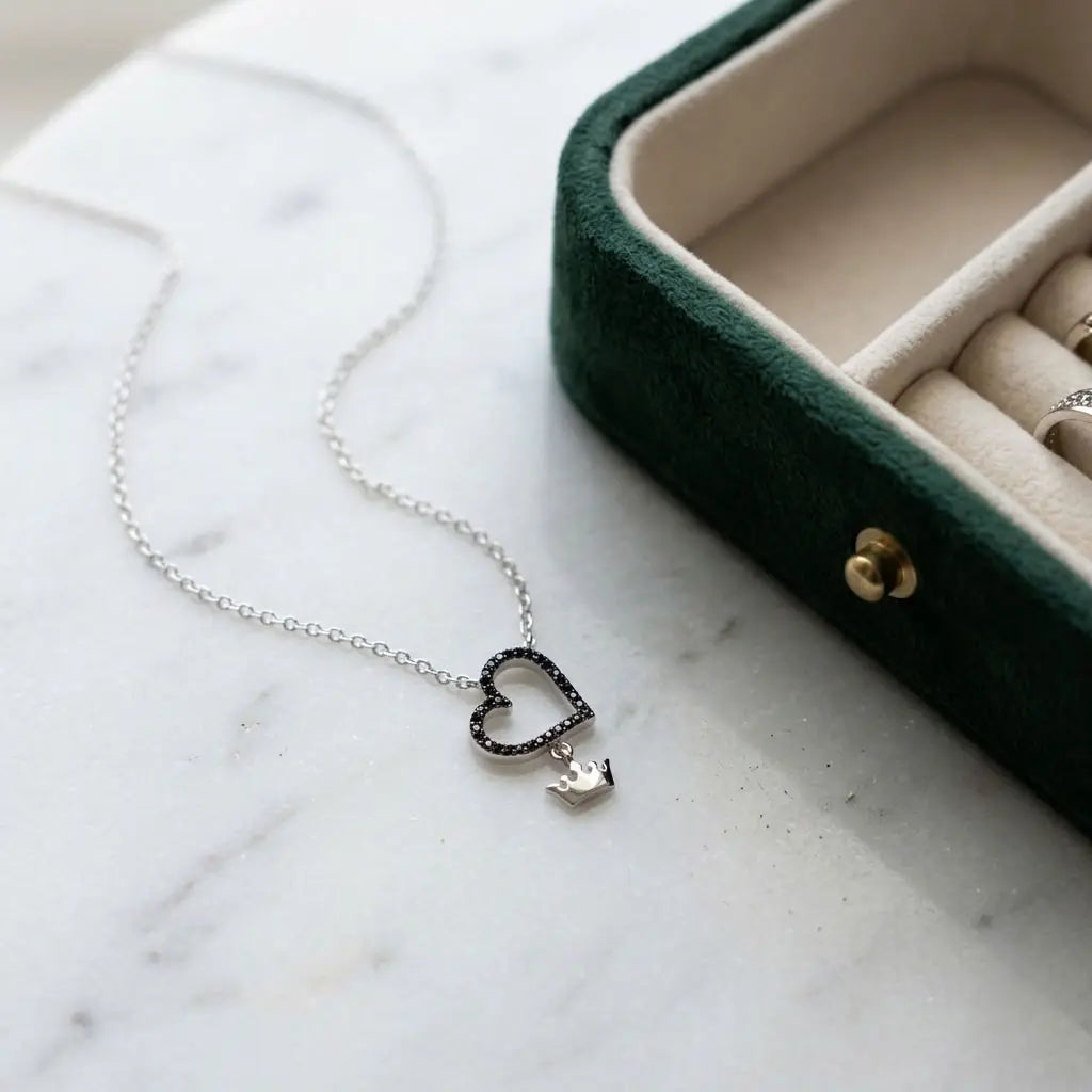 Black pavé open-heart necklace with polished silver crown charm on pale marble beside a green jewellery case.