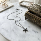Black pavé starfish pendant necklace lying on pale marble beside an ornate jewellery box, showing its curved five-arm design.