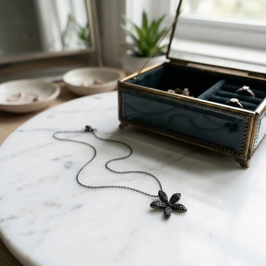 Black pavé flower pendant necklace on white marble beside an open jewellery box, showing the dark silver chain and five-petal design.