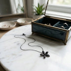 Black pavé flower pendant necklace on white marble beside an open jewellery box, showing the dark silver chain and five-petal design.