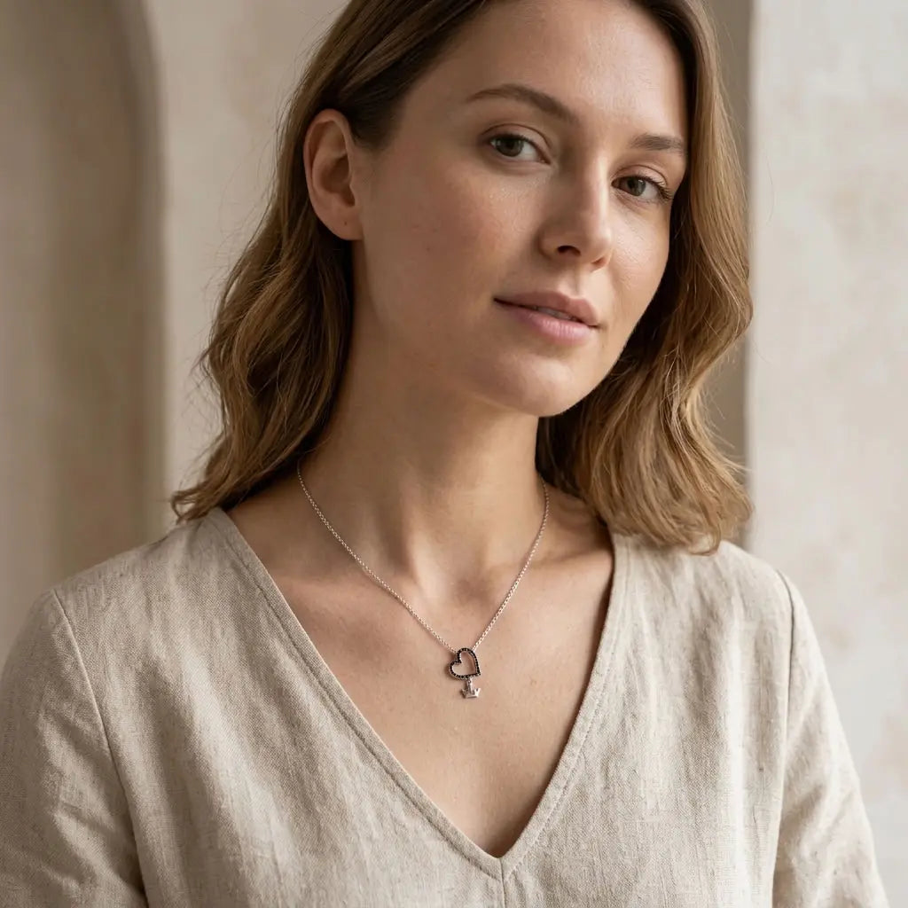 Model wearing a rhodium-plated silver necklace with black CZ open-heart pendant and small dangling crown over a light linen top.