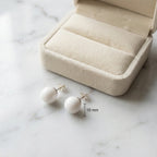 Pair of white sphere stud earrings in 925 sterling silver on white marble beside a cream jewellery box, with a 10mm size marker.