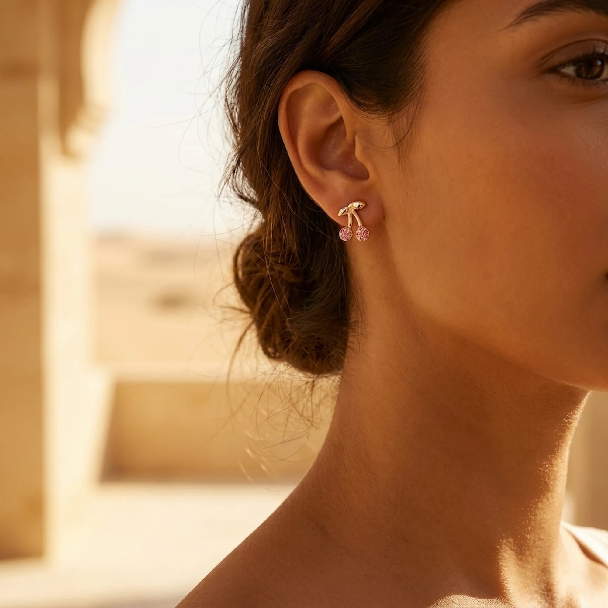 Cherry pavé stud earring worn on ear — warm 18K gold with rose crystals, Dubai heritage architecture background