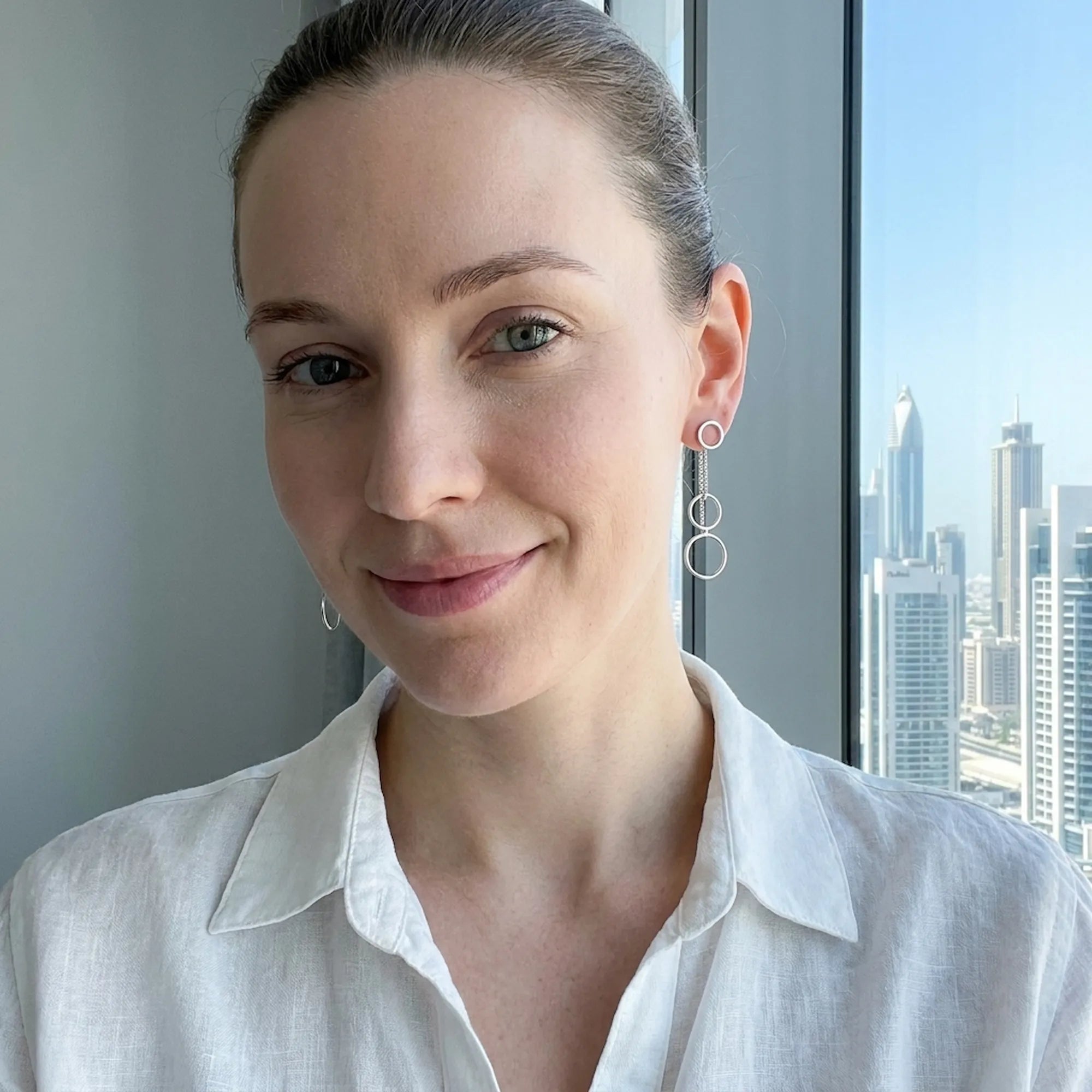 Woman wearing silver circle cascade earrings indoors at window with Sheikh Zayed Road Dubai skyline visible behind