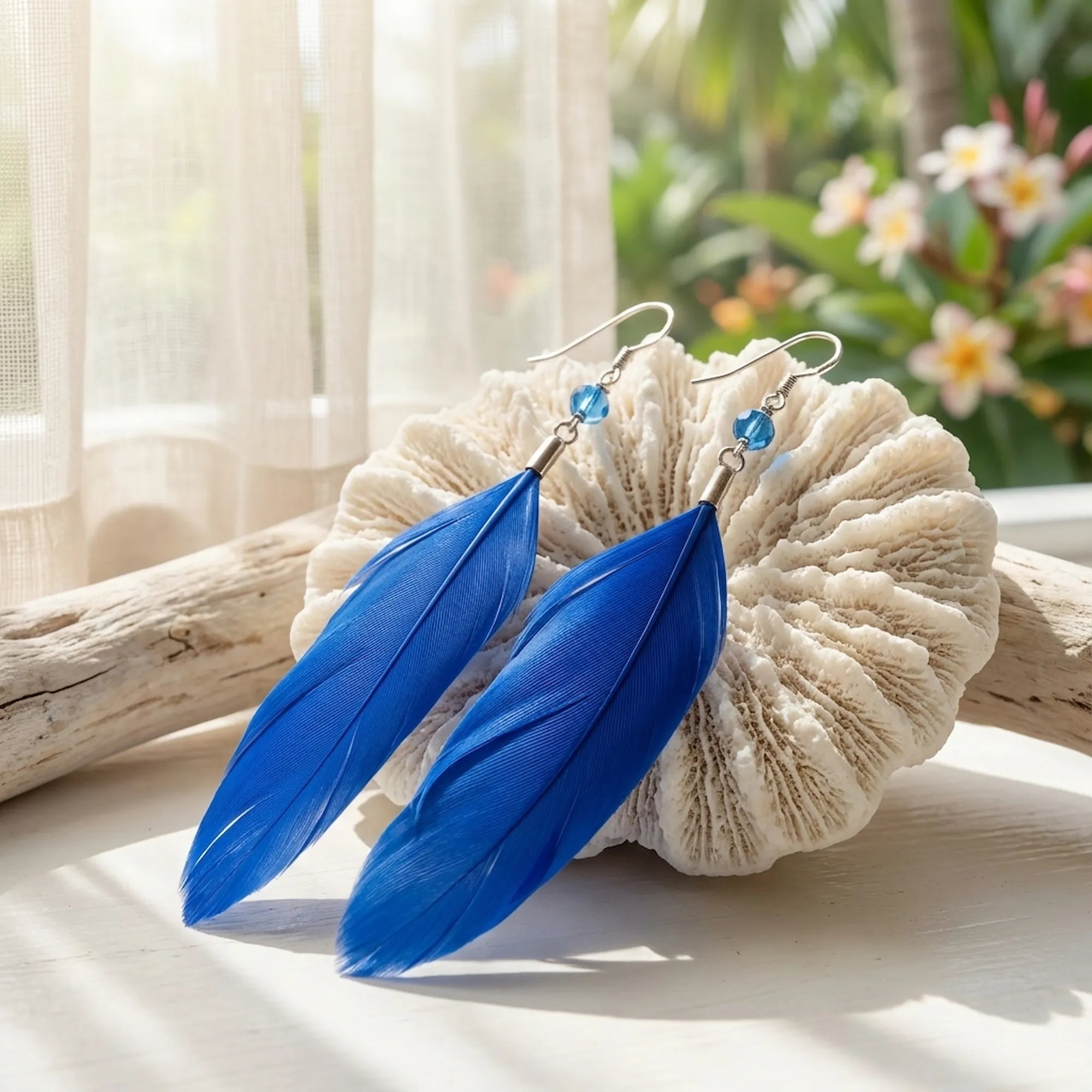 Cobalt blue feather earrings on white coral with tropical flowers and driftwood — natural light lifestyle flat lay Dubai