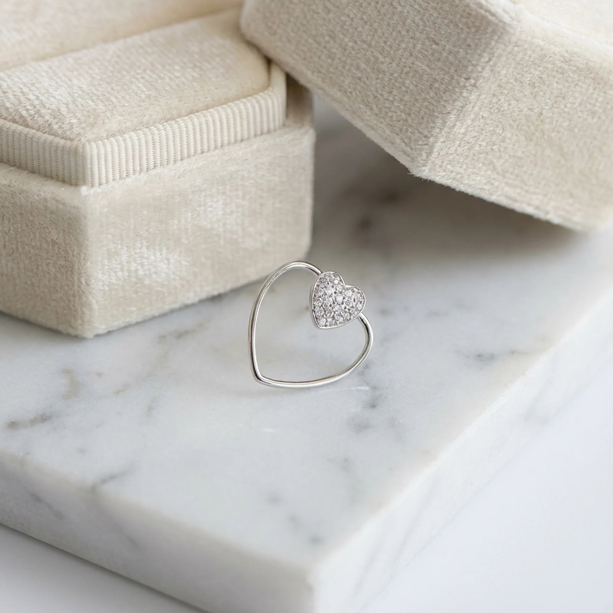 Pavé heart stud earring set within an open heart wire frame in sterling silver, on white marble beside a cream velvet box.