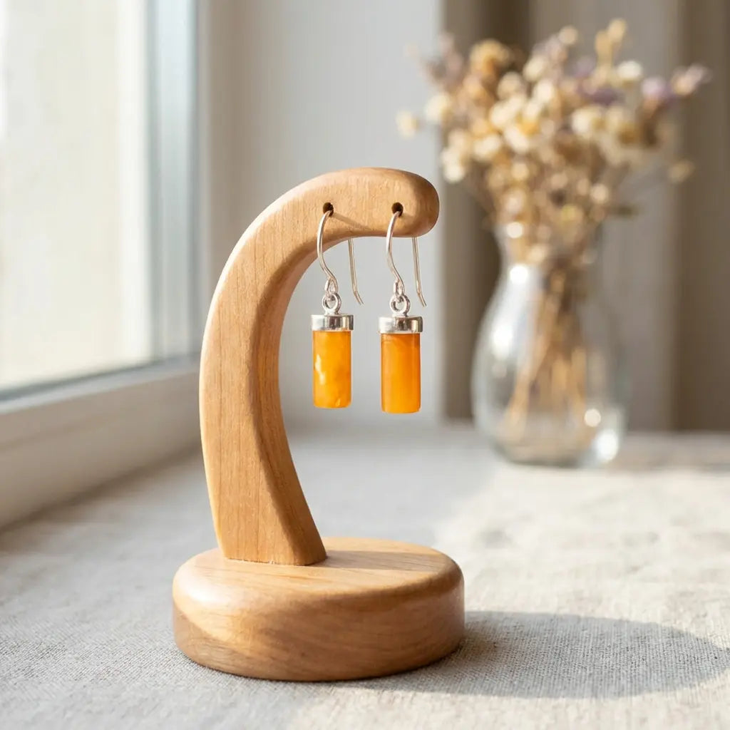 Cylindrical orange amber earrings hanging from a minimalist curved wooden jewelry display stand near a bright window with a floral vase