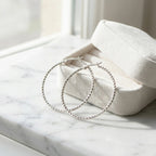 Large 925 silver diamond-cut hoop earrings resting in a cream velvet gift box on marble. Luna Dura.
