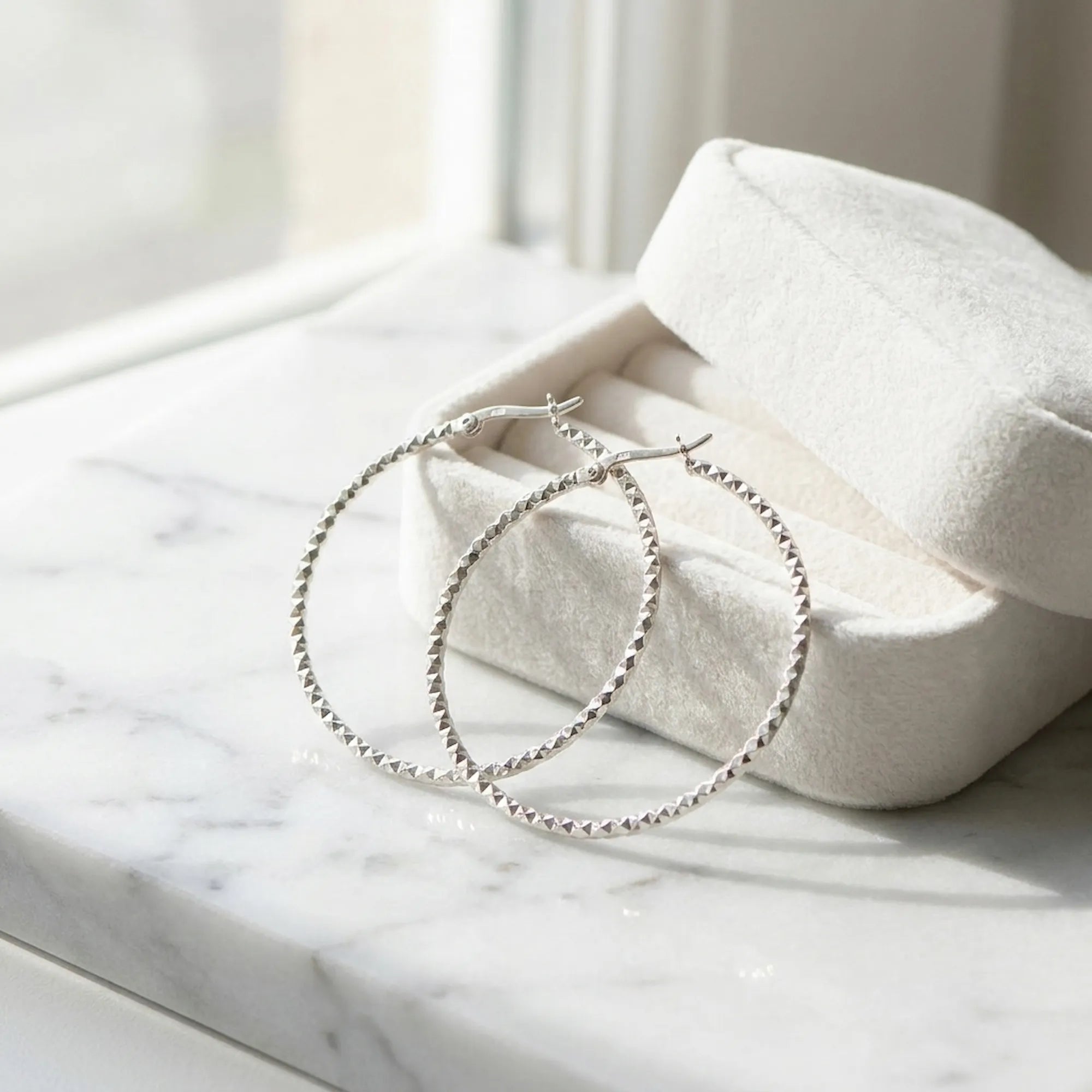 Large 925 silver diamond-cut hoop earrings resting in a cream velvet gift box on marble. Luna Dura.