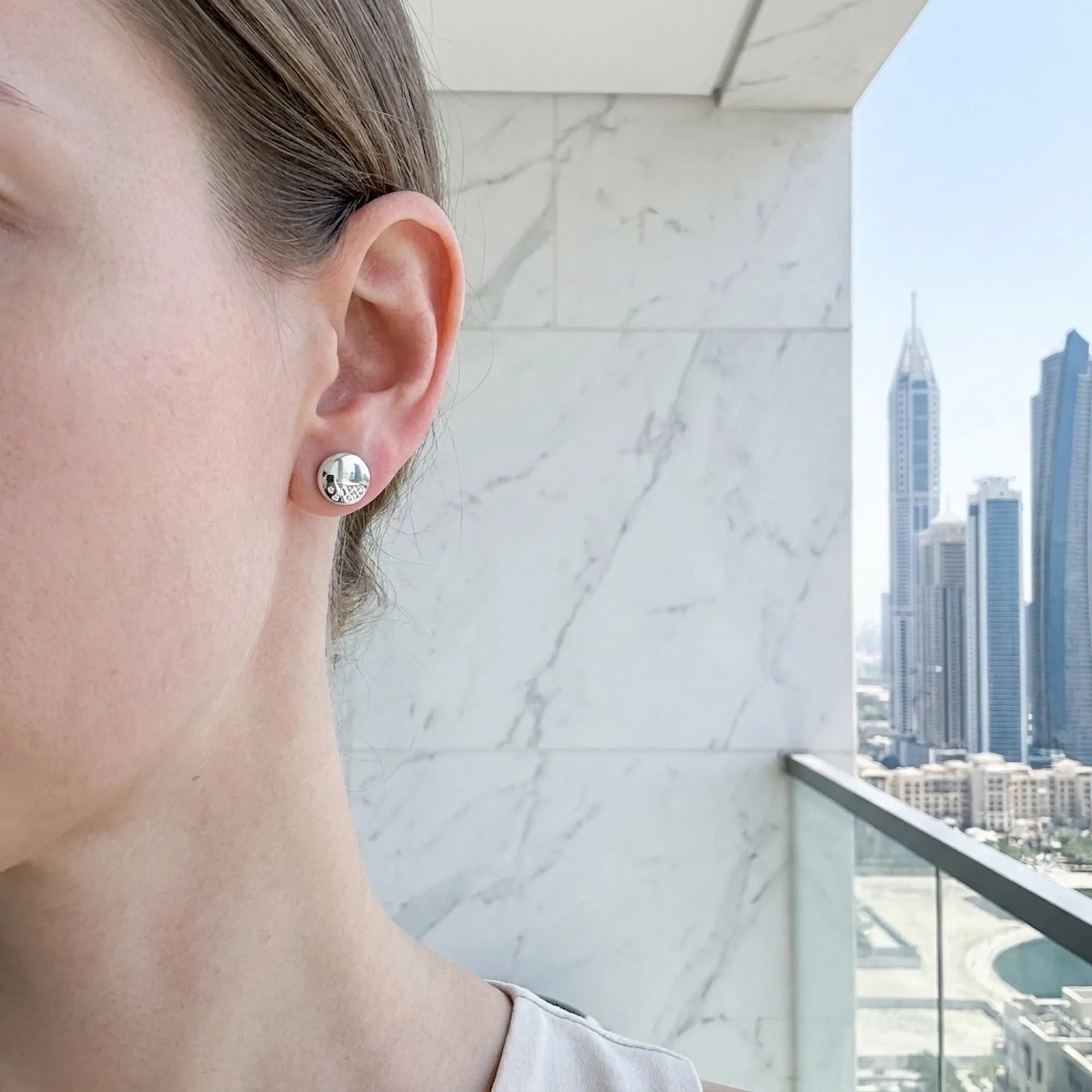 Round dome stud earring on model's ear with Dubai skyline skyscrapers in background — 925 sterling silver