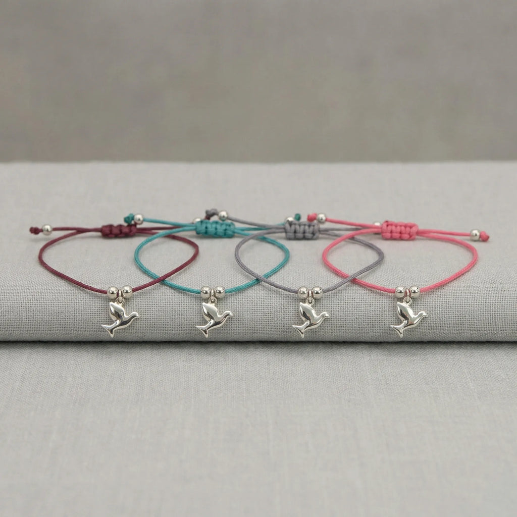 All four silver dove charm bracelets laid on grey linen showing burgundy, teal blue, lavender and rose pink cord options UAE.