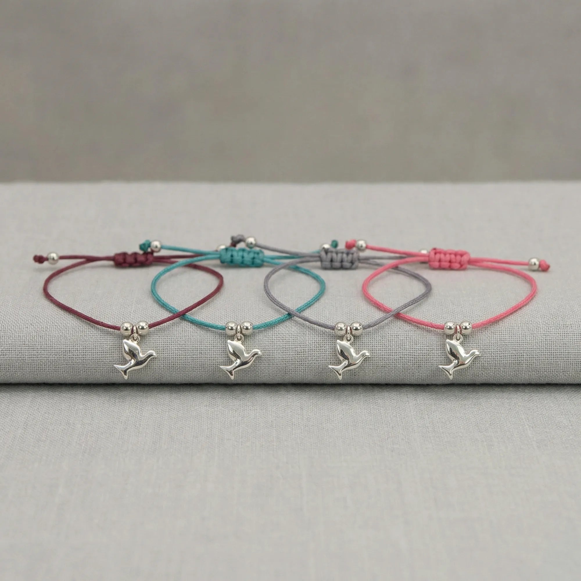 All four silver dove charm bracelets laid on grey linen showing burgundy, teal blue, lavender and rose pink cord options UAE.