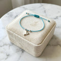 Silver dove charm bracelet on teal blue cord over closed velvet gift box on marble surface in soft light