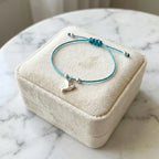 Silver dove charm bracelet on teal blue cord over closed velvet gift box on marble surface in soft light