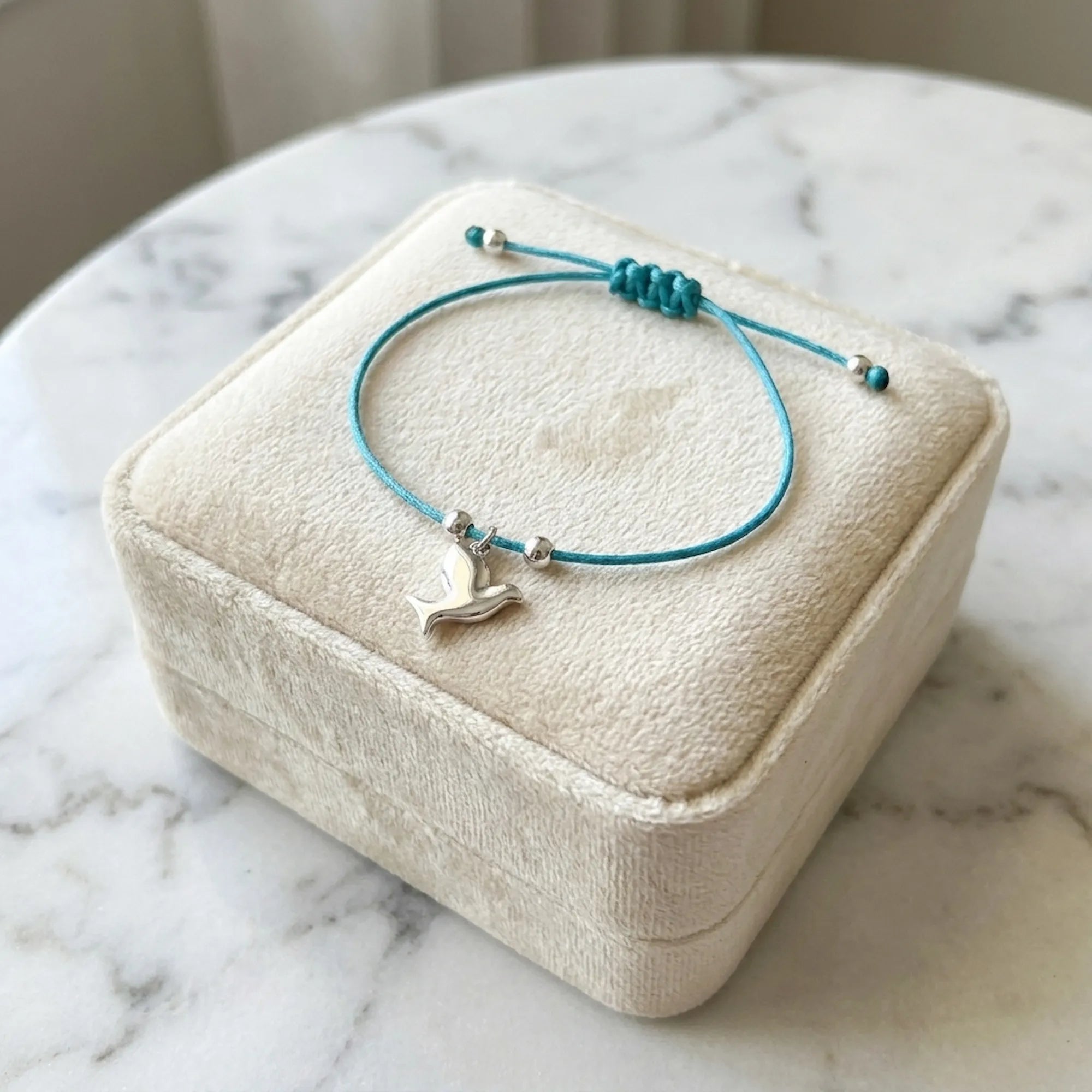 Silver dove charm bracelet on teal blue cord over closed velvet gift box on marble surface in soft light