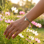 Silver dove charm bracelet on teal blue cord worn on wrist with hand extended among pink and white wildflowers