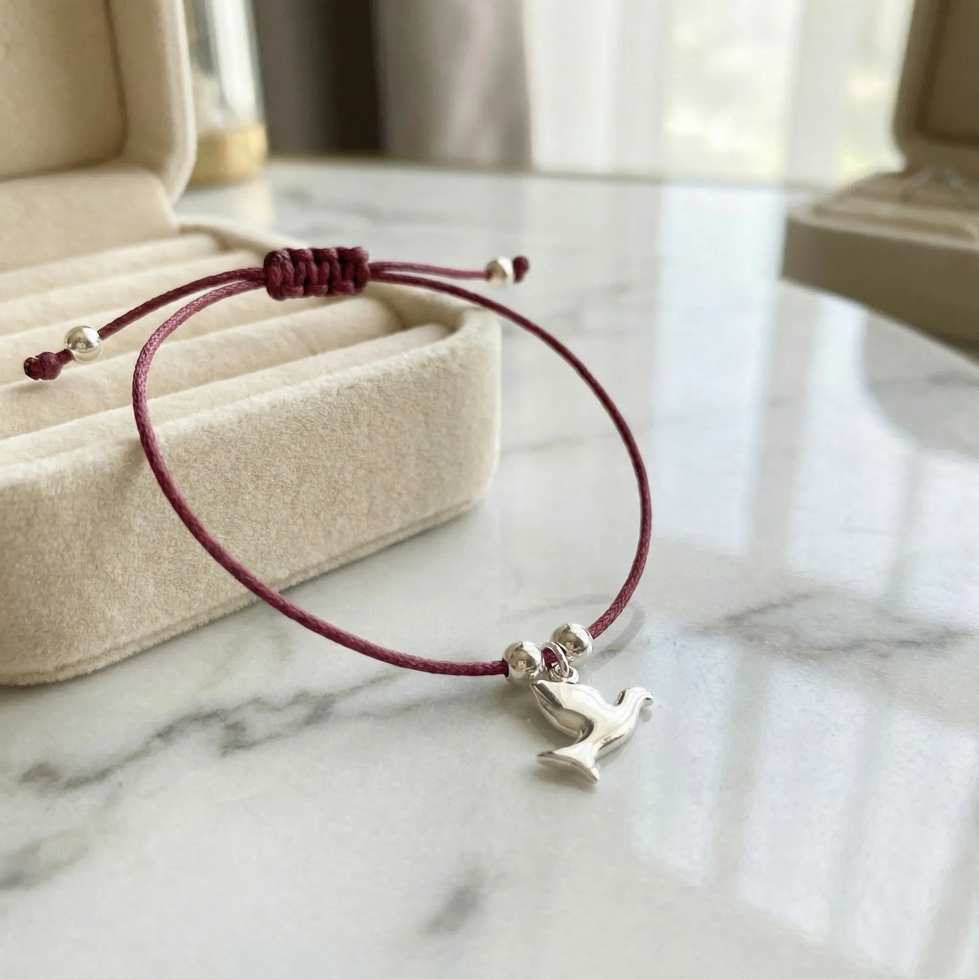 Burgundy cord silver dove charm bracelet laid flat on white marble surface beside open cream velvet jewellery tray