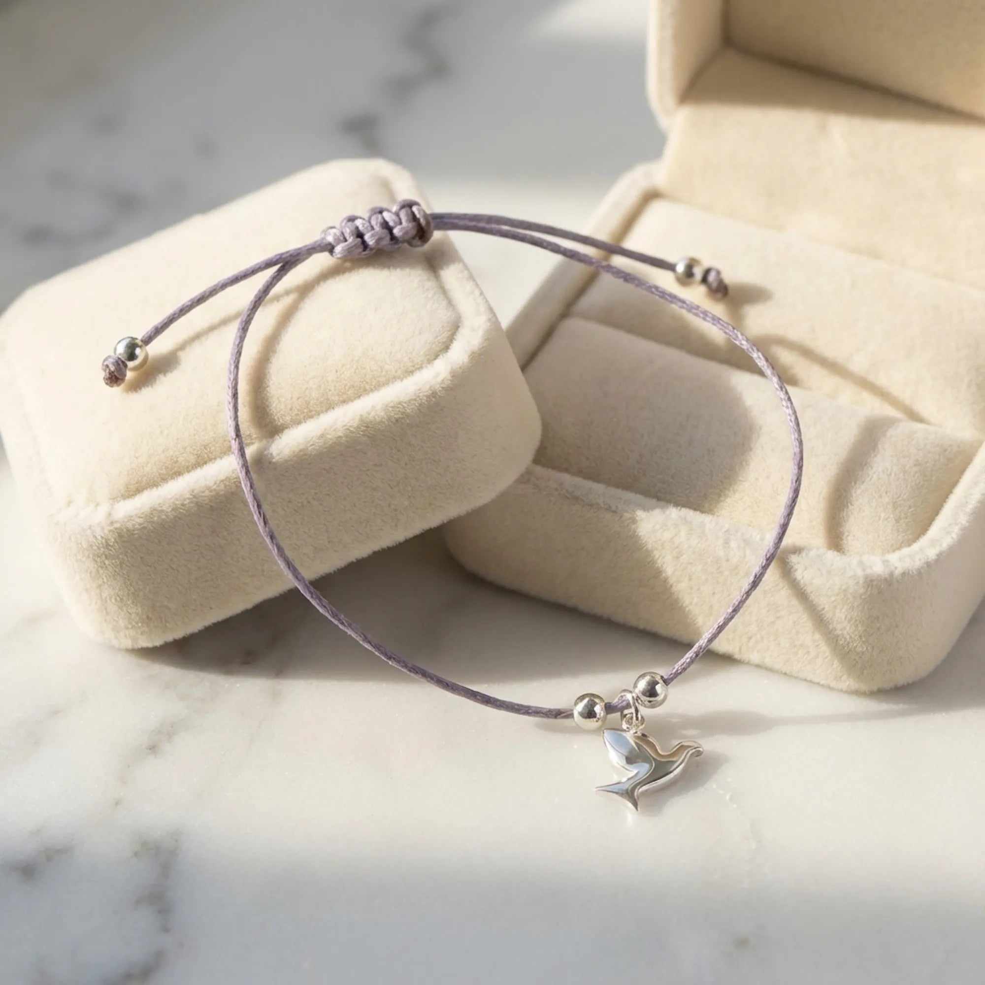 Lavender cord silver dove charm bracelet draped over open Luna Dura cream velvet gift box on white marble surface