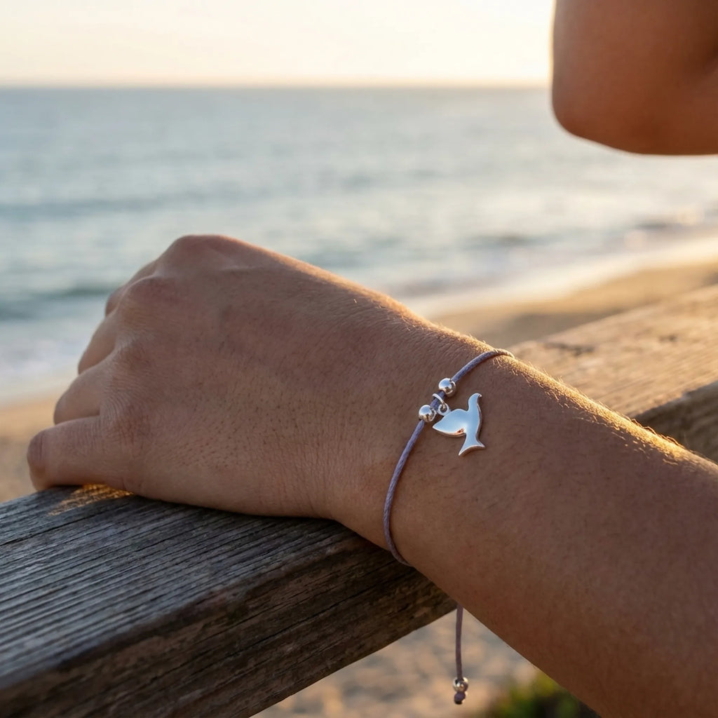 Silver dove charm bracelet on lavender cord worn on wrist at golden hour with La Mer Jumeirah beach behind