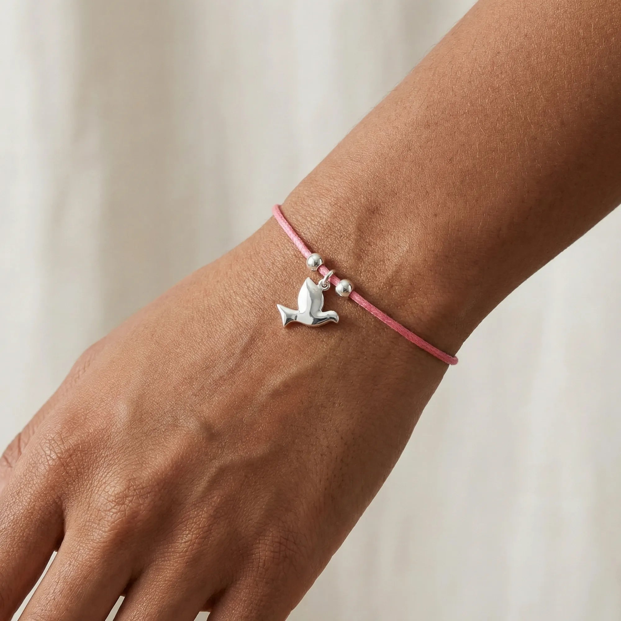 Silver dove charm bracelet on rose pink cord worn on wrist showing polished mid-flight dove charm and silver beads