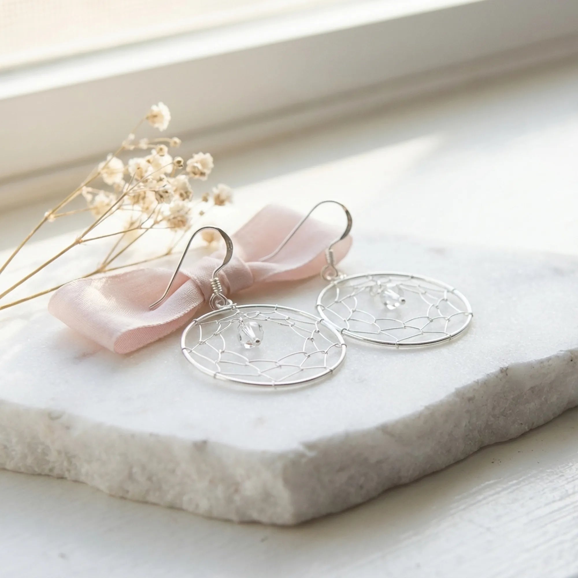 Silver dreamcatcher drop earrings on white stone surface with dried flowers and pink ribbon in soft natural light UAE