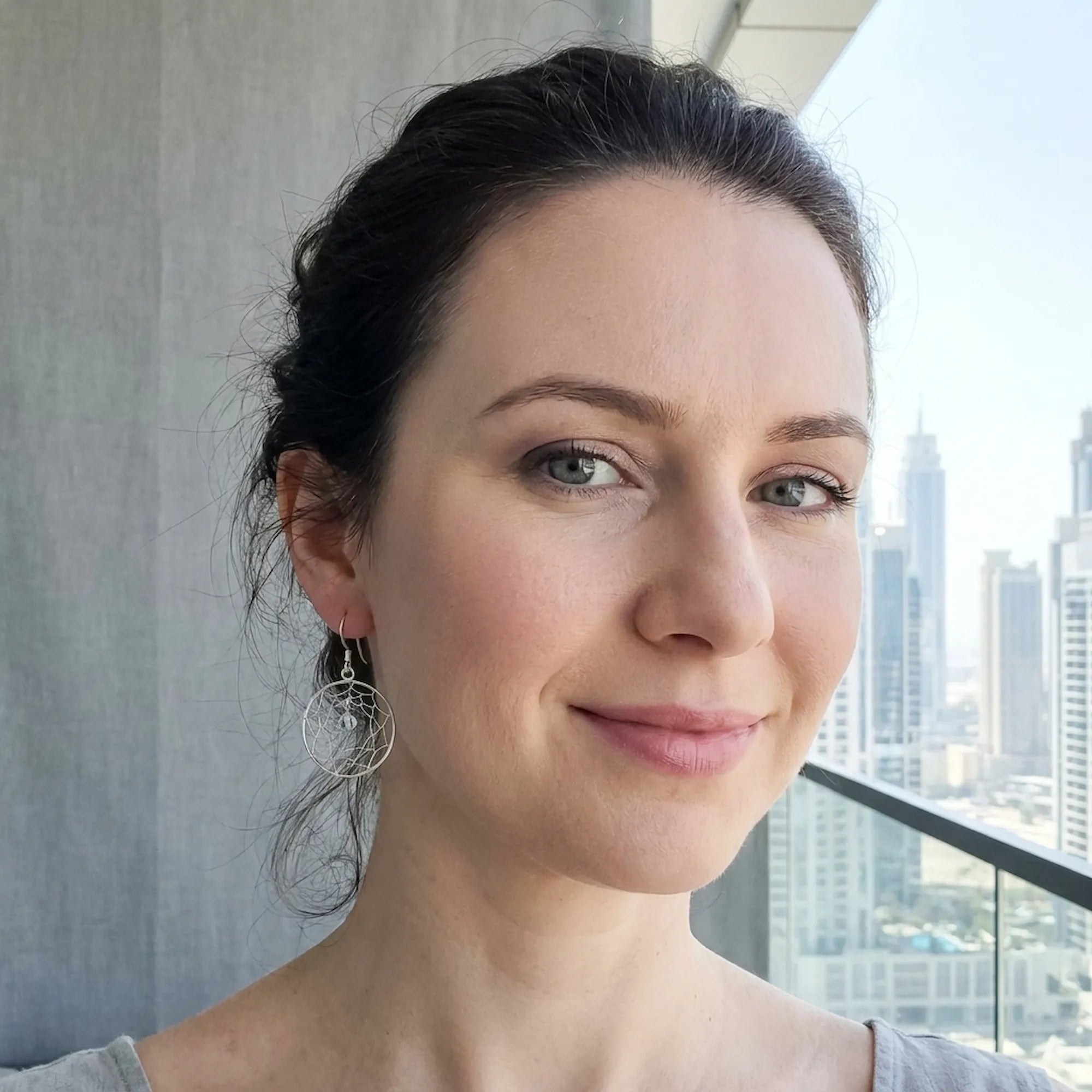 Woman wearing silver dreamcatcher drop earrings indoors with Dubai city skyline visible through window behind her