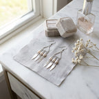 Silver geometric pearl drop earrings resting on a grey cloth next to a beige velvet ring box and glass perfume bottle.