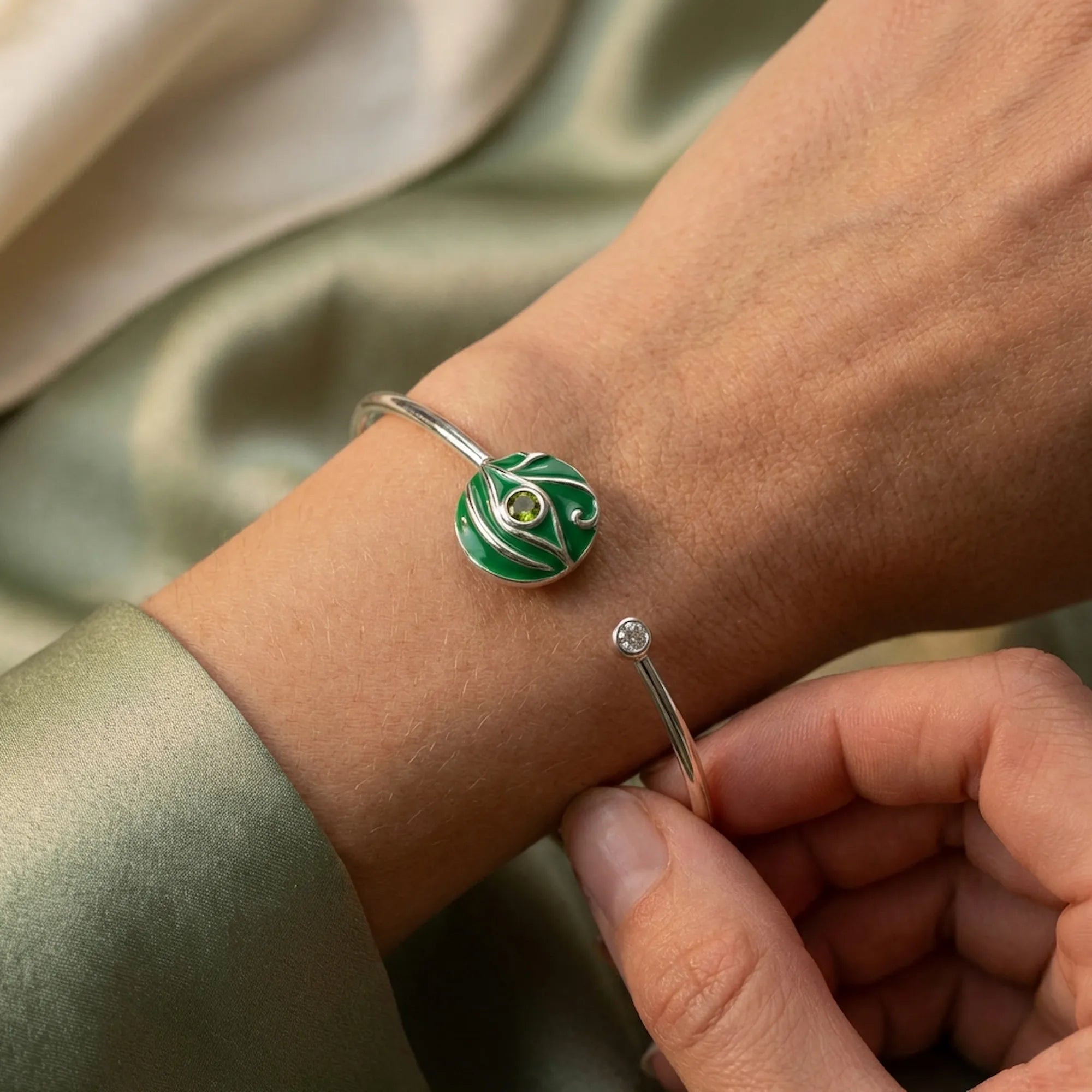 Close-up of green peridot evil eye cuff bangle being adjusted on wrist, sage green fabric background
