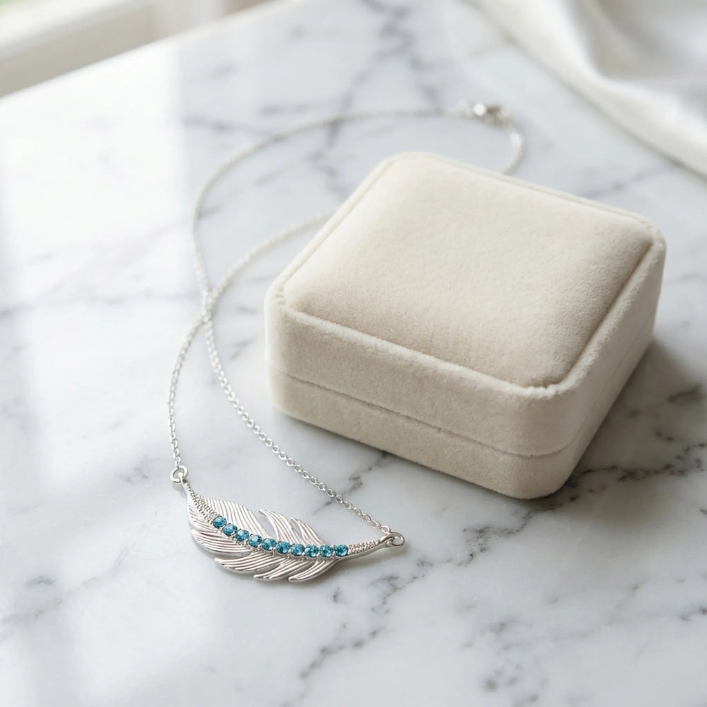 Silver feather necklace with turquoise crystal beside a cream velvet gift box on marble, Luna Dura Dubai.