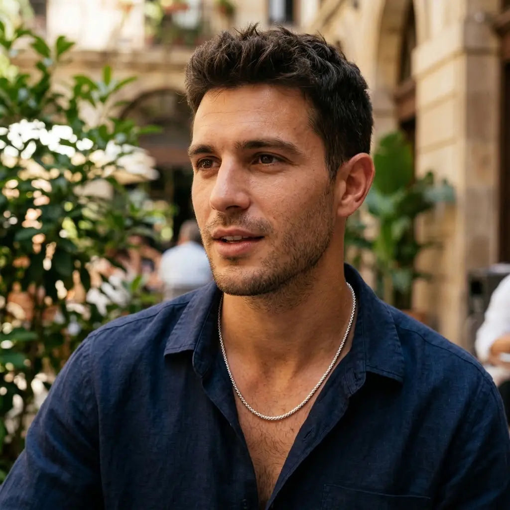 Man wearing a fine silver-tone necklace with an open-collar navy shirt outdoors.