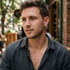 Man wearing a long, narrow silver link chain necklace with a dark open-collar shirt.