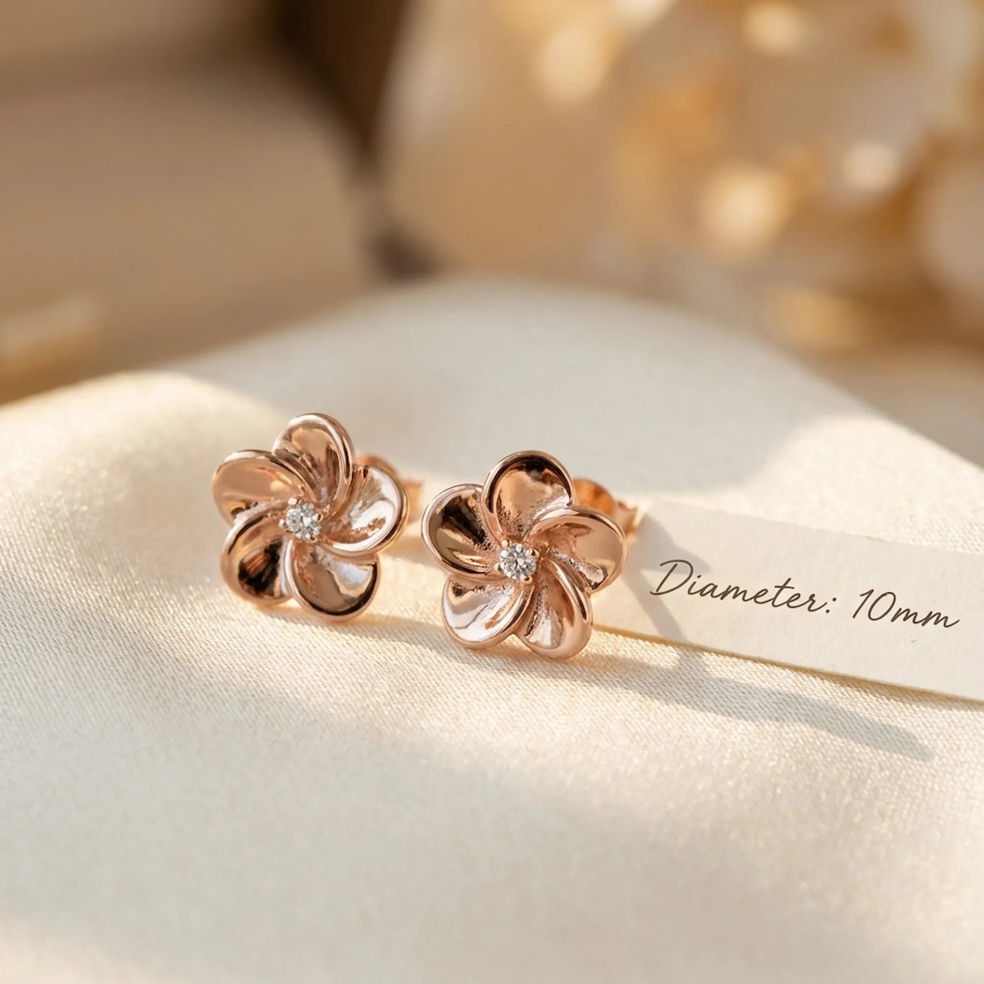 Pair of rose gold flower stud earrings on cream linen with 10 mm diameter measurement detail