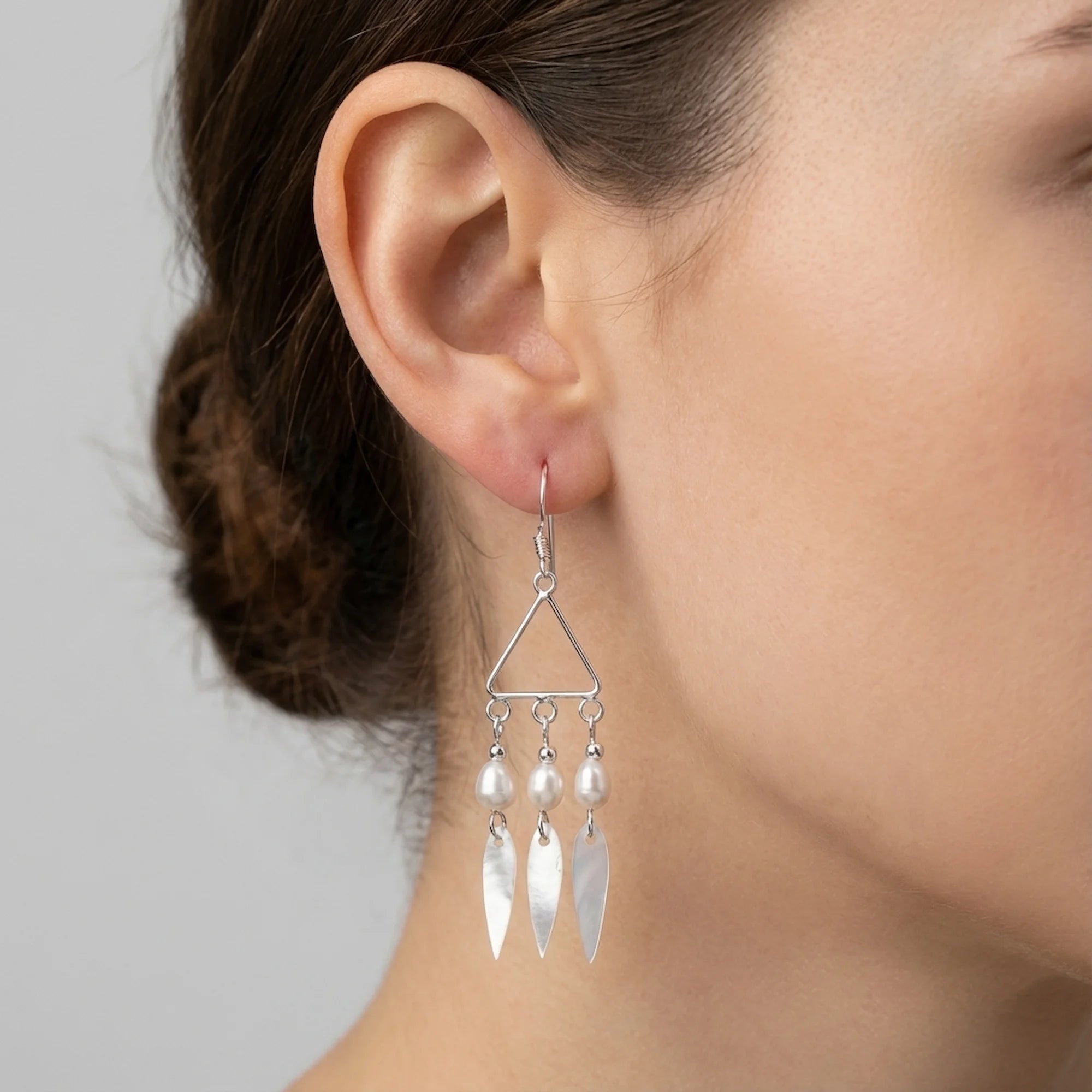 Woman wearing geometric triangle silver drop earrings with pearl and shell accents, facing right against a grey wall.
