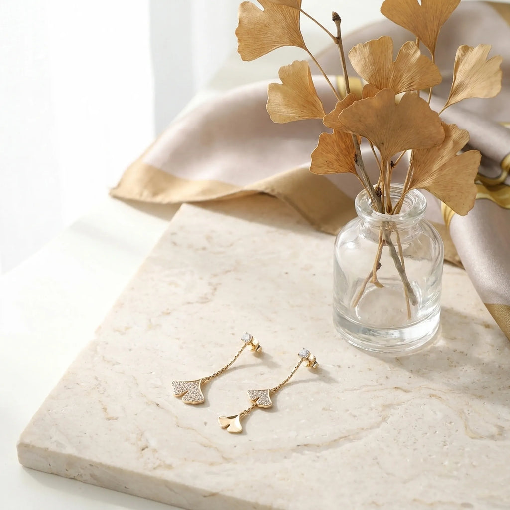 Gold ginkgo drop earrings on marble surface with dried ginkgo leaf branch in glass vase, flat lay