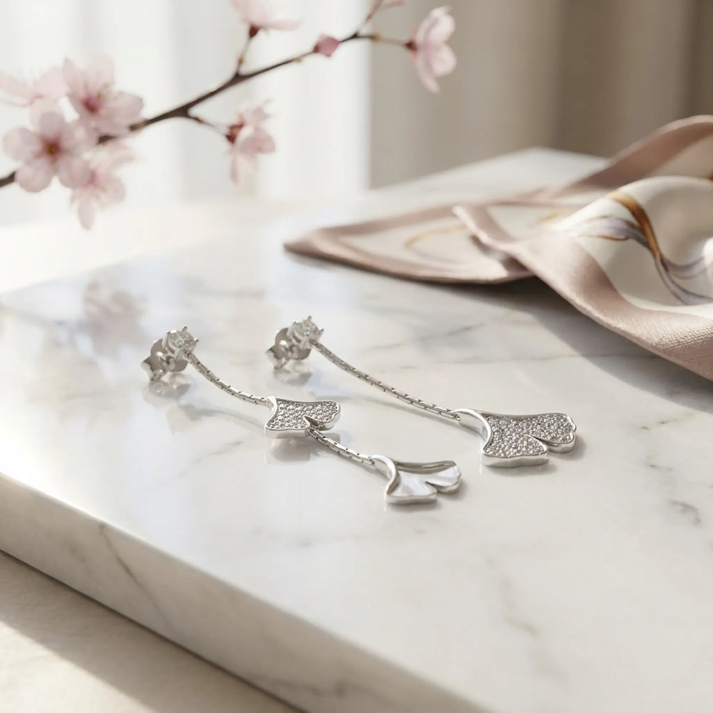 Rhodium silver ginkgo drop earrings on white marble with cherry blossom branch, styled flat lay