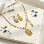 Gold Hamsa necklace and earrings flatlay on travertine with lavender and blue stones, gift styling, Dubai