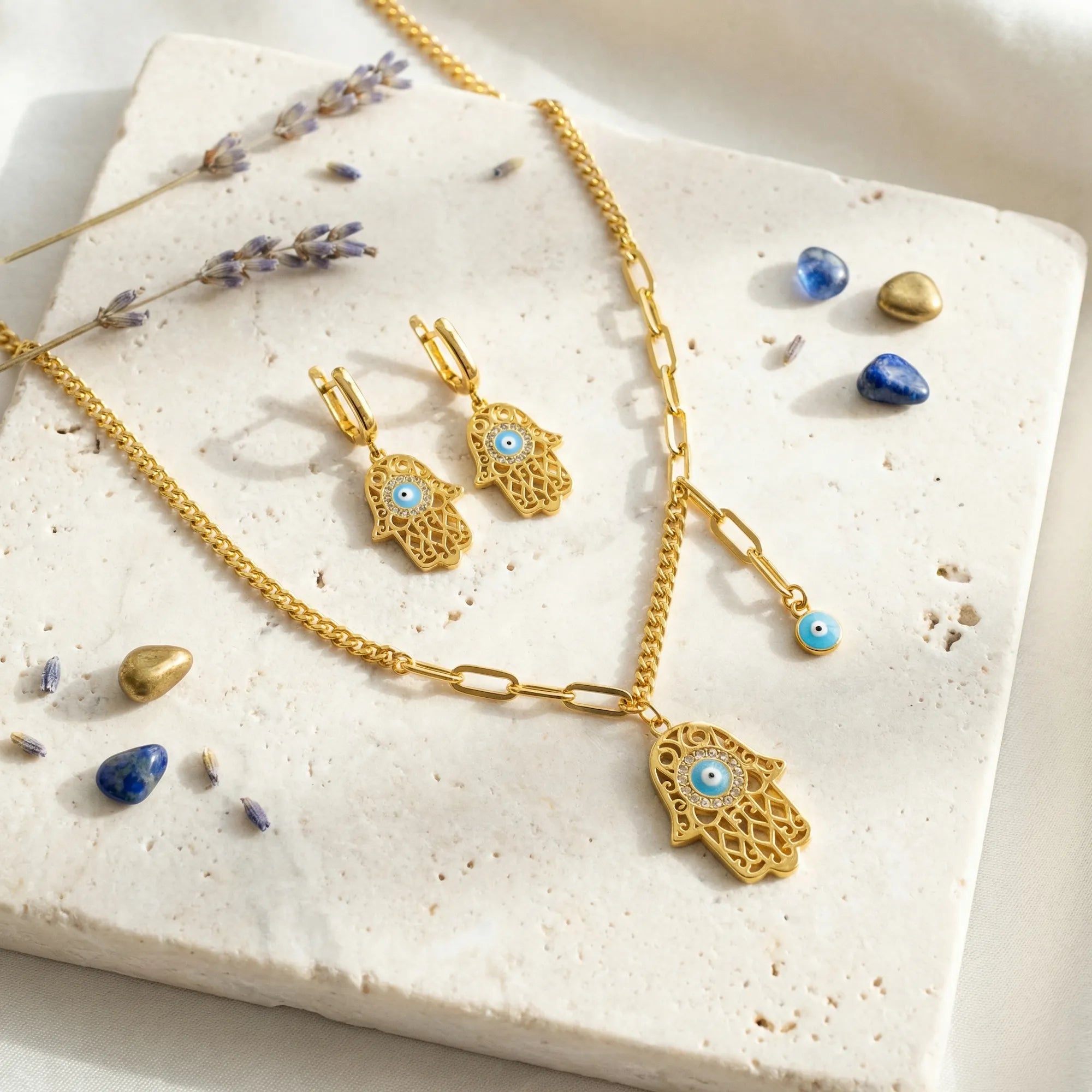 Gold Hamsa necklace and earrings flatlay on travertine with lavender and blue stones, gift styling, Dubai
