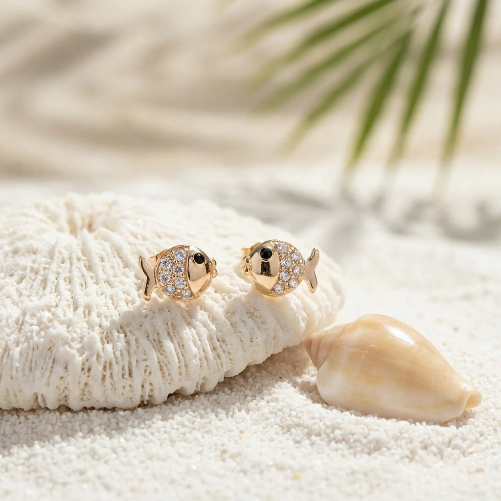 Goldfish stud earrings on white coral and shell in sand — gold 925 silver, beach flat lay with palm leaf