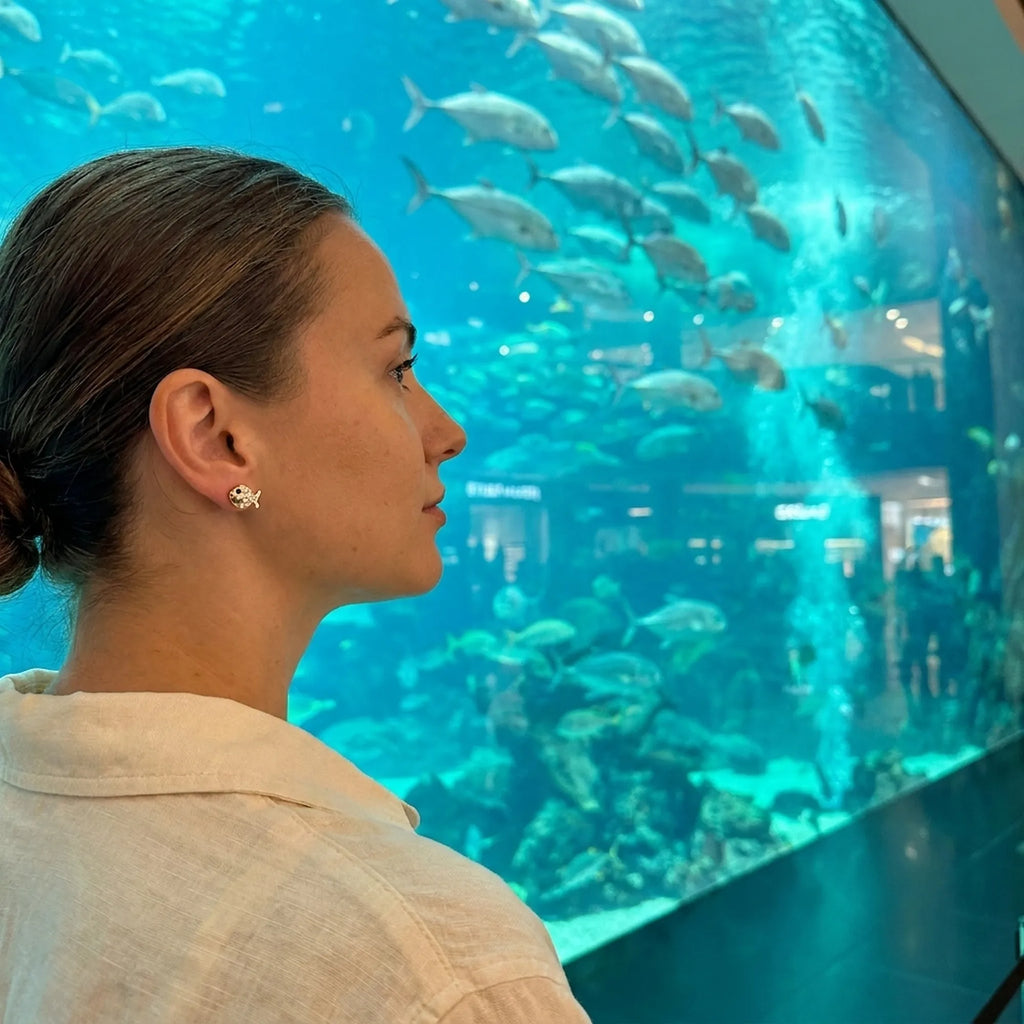 Goldfish stud earrings at Dubai Aquarium — gold 925 silver CZ stud, sharks and fish in background, second lifestyle