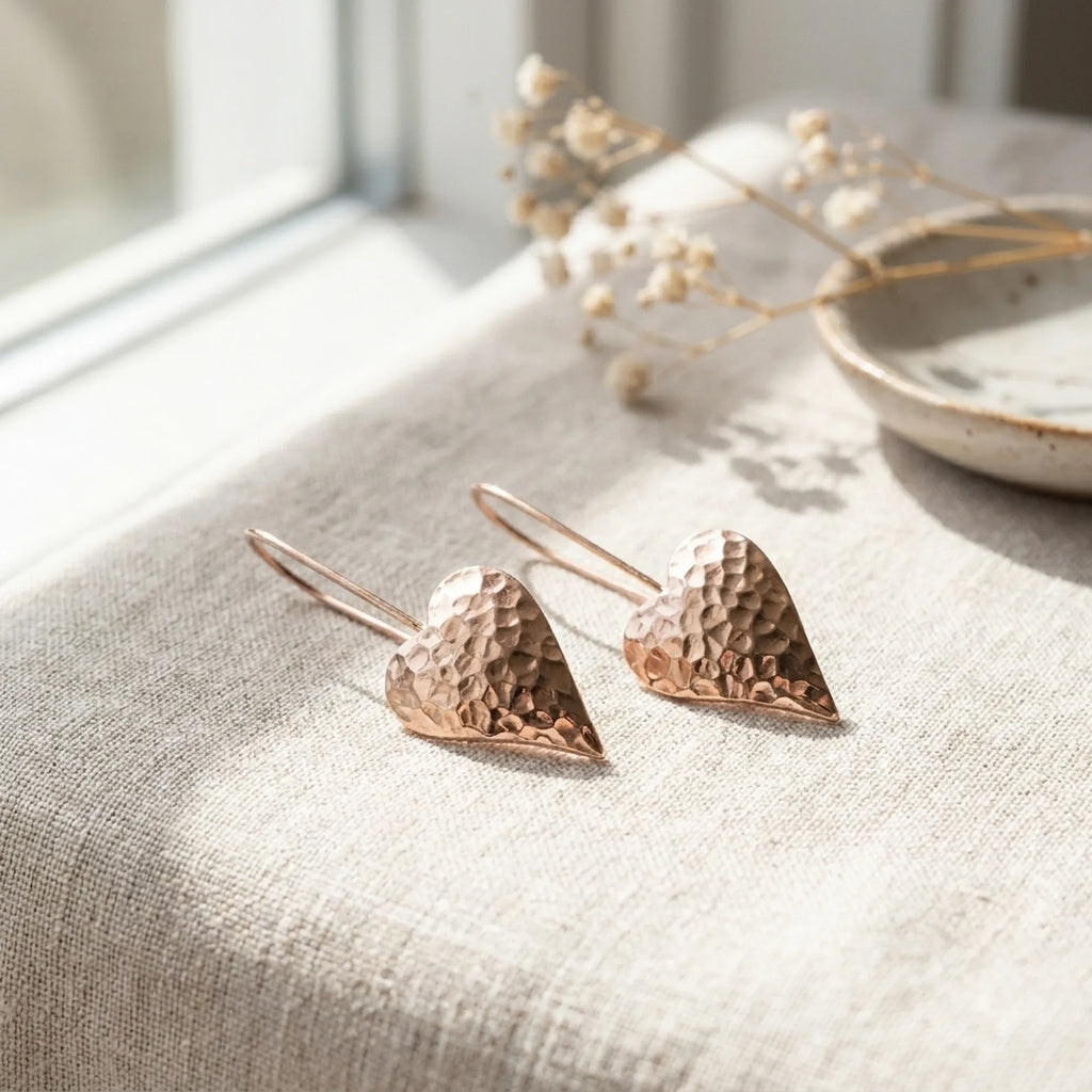 Hammered rose gold heart drop earrings on linen with dried gypsophila — natural light flat lay showing faceted surface and tapered heart form