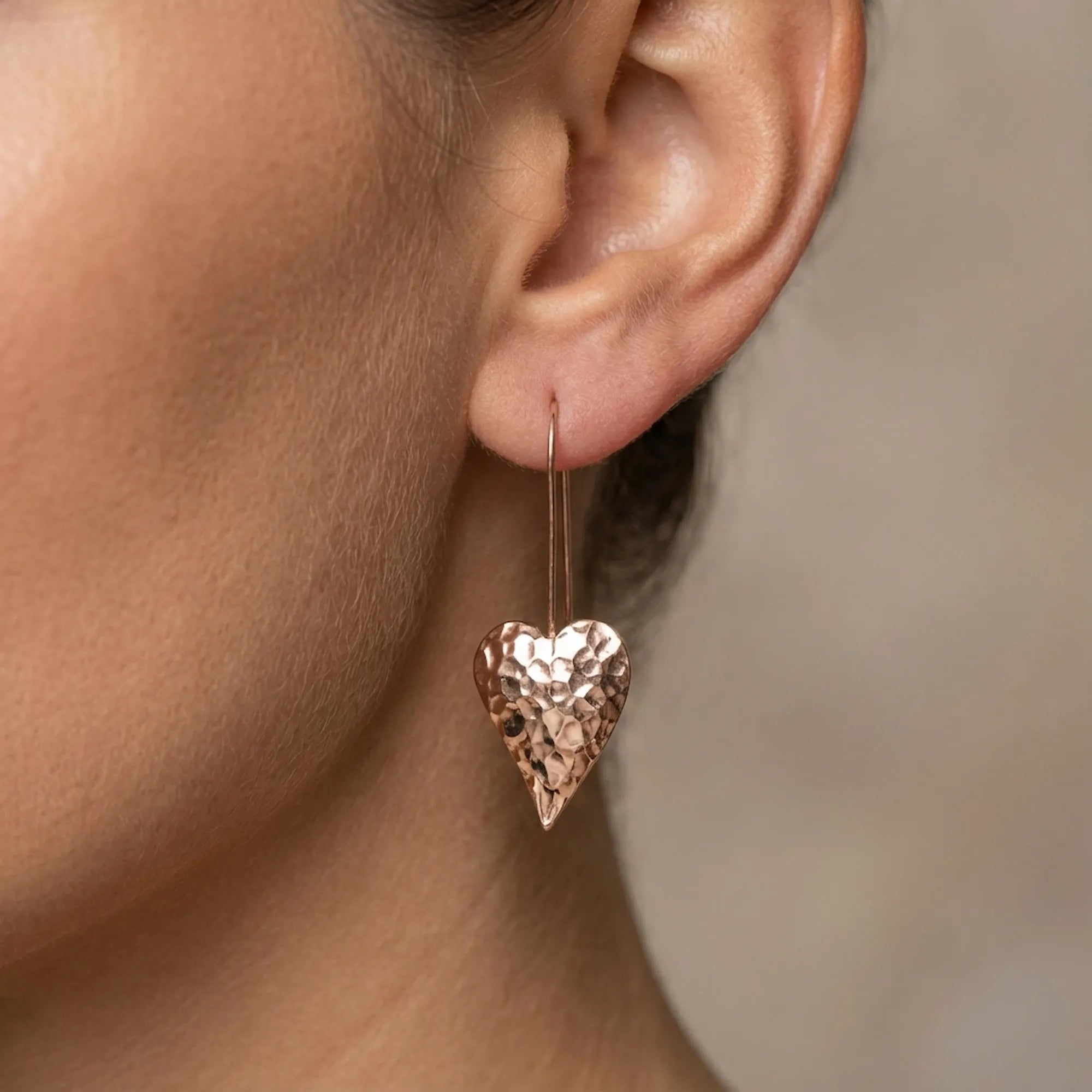 Close-up of hammered rose gold heart drop earring on ear — faceted surface detail and wire hook, 44mm elongated tapered heart