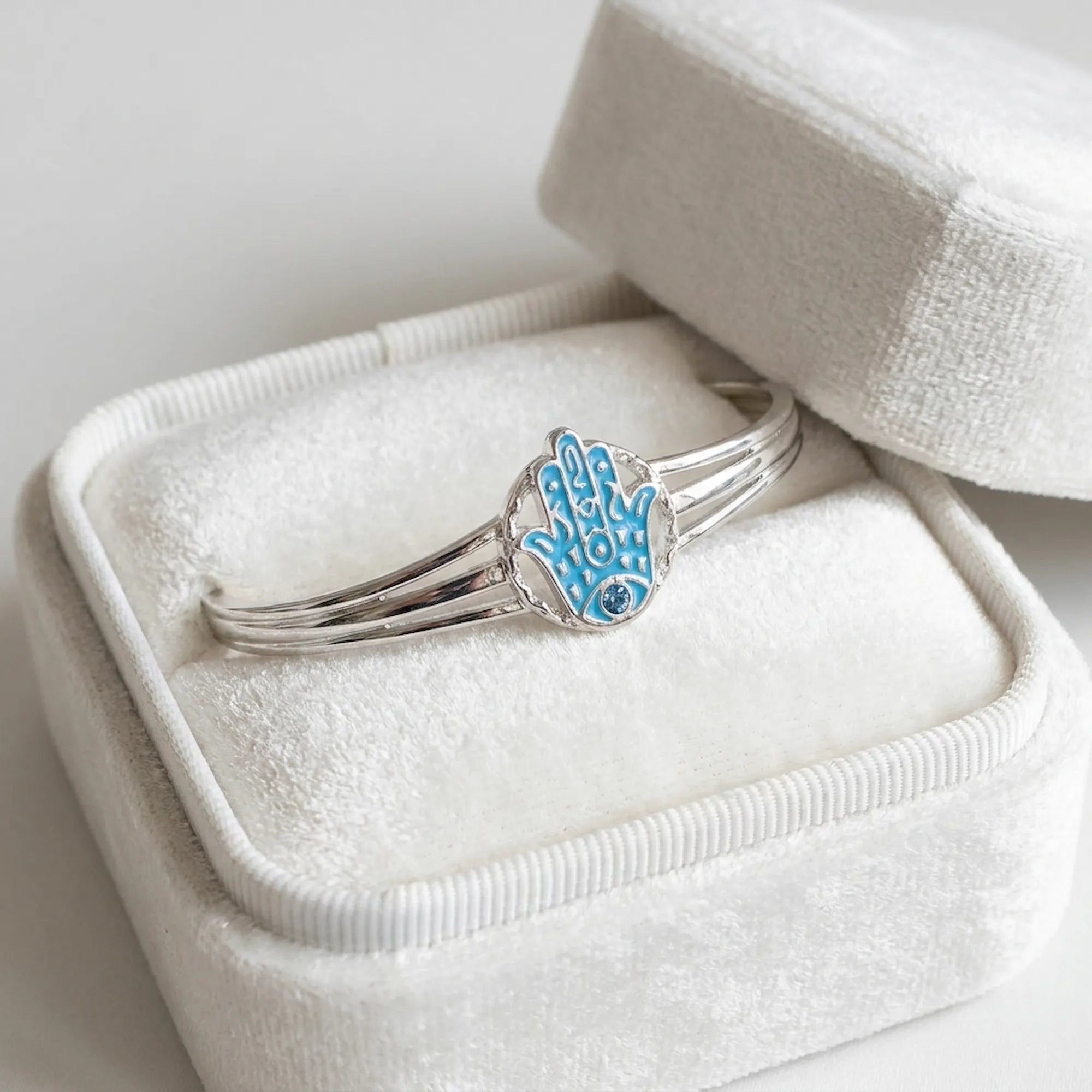 Powder blue enamel Hamsa bangle resting in open cream velvet gift box, sky blue topaz eye visible